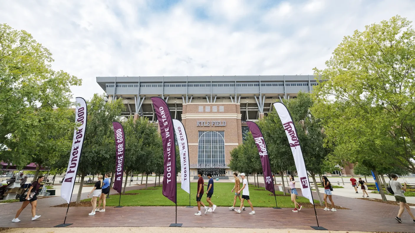 Students walking in between the "Together, we stand as a force for good" and "Howdy" feather banners in front of Kyle Field
