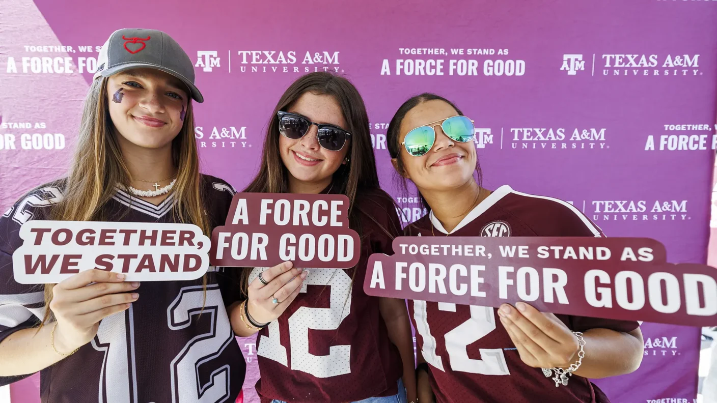 Three aggie football fans standing in front of a branded Texas A&M backdrop holding signs supporting the "Together, we stand as a force for good" campaign