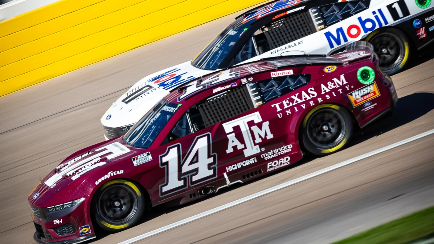 Two NASCAR vehicles racing on a track, with the Texas A&M branded #14 car in the lead