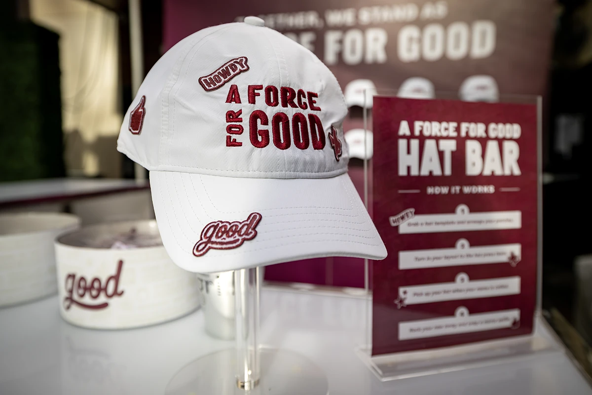A white A Force for Good hat covered with Texas A&M brand patches sits on a stand next to a sign that says "A Force for Good Hat Bar"