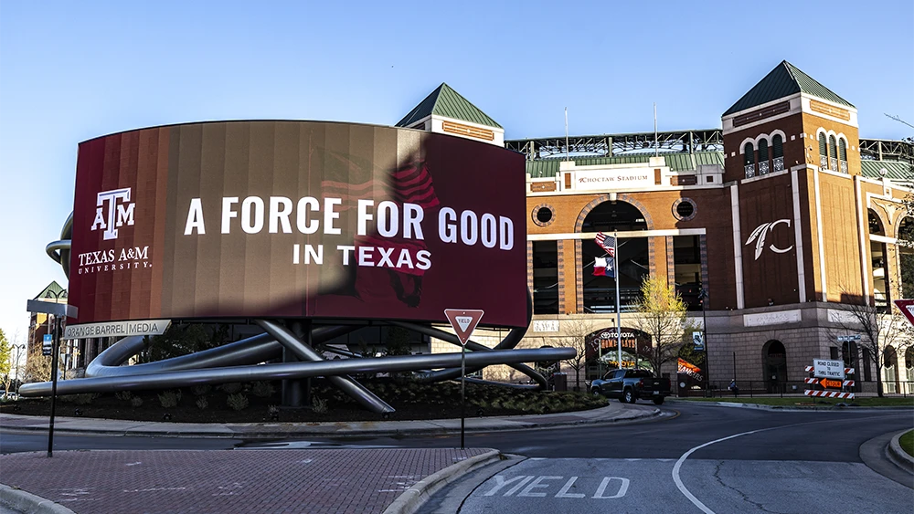 A round electronic billboard outside of the race says "A Force for good in Texas" with the Texas A&M logo